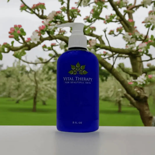 Vital Therapy Apple Delight Cleanser bottle in front of a blooming tree, offers soy-free, non-GMO skincare benefits.